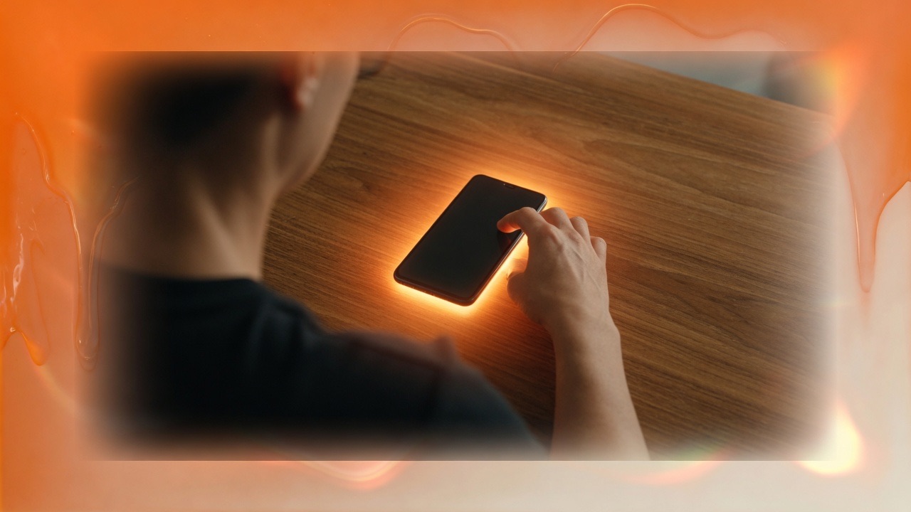An adult at a wooden desk with a glowing phone, framed in peach glass